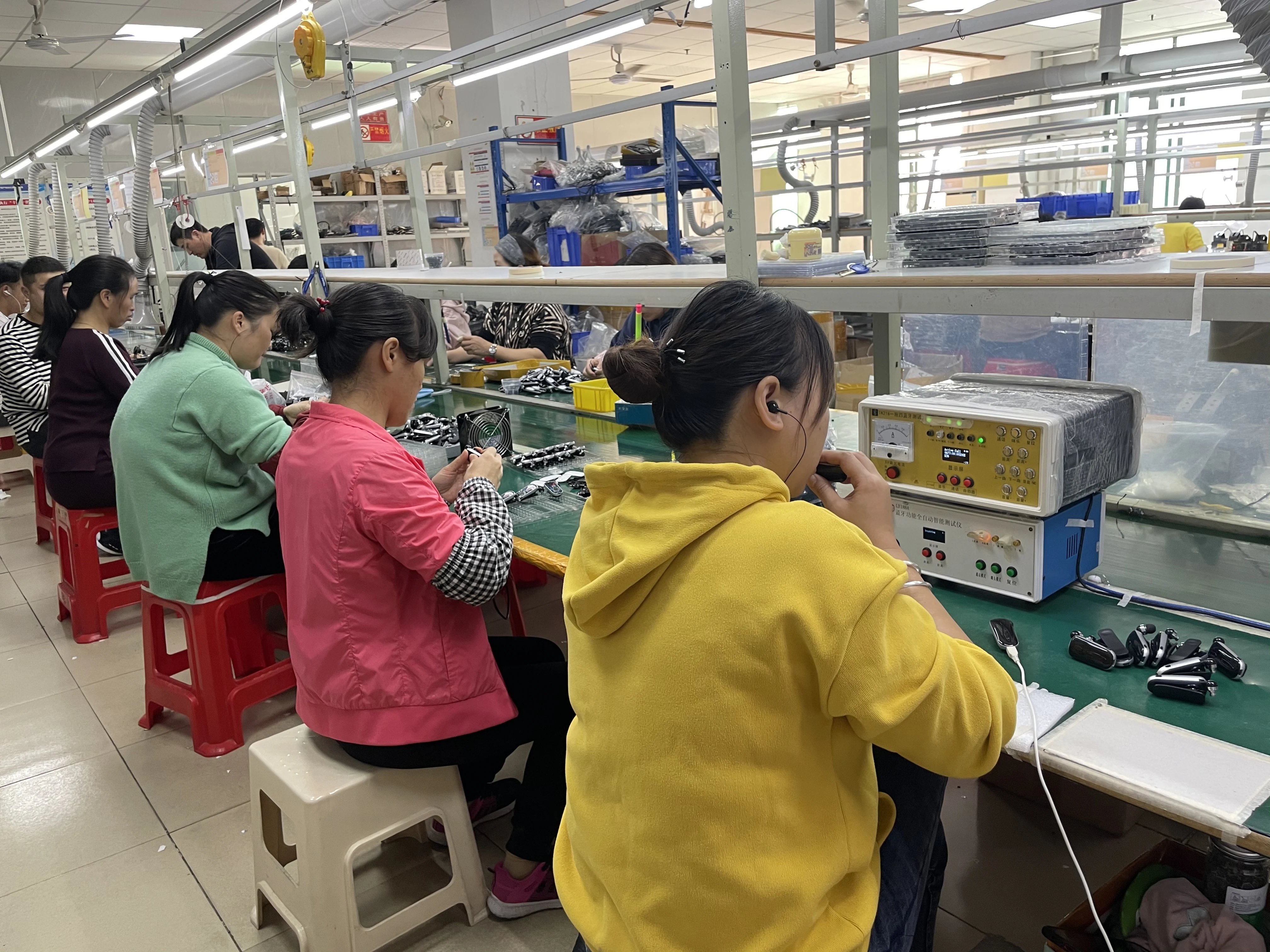 Bench lighting, tools and containers are positioned around seated operators. The close-up makes the working area easier to understand than a wide factory photograph alone: individual tasks take place at the bench, while shelves above the line hold materials within reach. This view shows the practical layout used for hands-on earphone production.